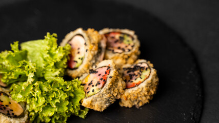 Custom sushi roll in tempura with nori, fresh salmon, tuna, avocado, masago caviar, drizzled with pineapple sauce with salad pouring as decoration on a black plate on a black table and background.