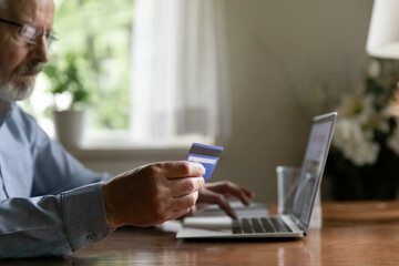 Close up focused mature man making secure internet payment, holding plastic credit or debit card, entering information, using laptop, shopping or checking balance online, paying bills, purchasing