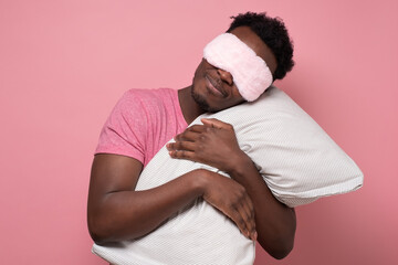 African young man holding a white pillow, sleeping. Studio shot on pink wall.