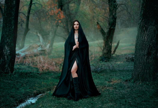 Mysterious fantasy gothic woman dark witch obsessed by evil. Girl demon vampire in black dress cape hood. walk in dark dense deep forest background, trees. Medieval queen in silk cloak, scarf posing