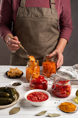 The concept of fermented canned vegetarian food. Carrots, tomatoes, cucumbers, pickled, sauerkraut sour in glass jars and plates on a white wooden kitchen table. Home-made preparations.