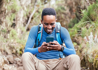 Young African ethnicity man video chatting via cellphone with somebody and cheerfully laughing as he having a hiking walk in the forest. Happy people, network technology, or traveling concept