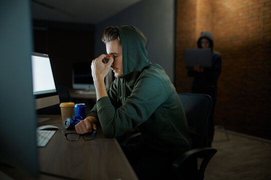 Tired male hacker in hood sitting at monitors
