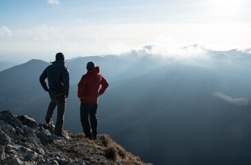 Two men standing standing with trekking poles on cliff edge and looking at sunset rays over the clouds. Successful summit concept image.