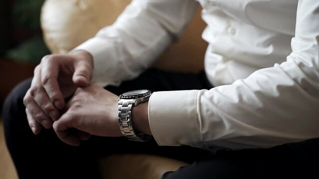 A stylish guy with a watch on his hands is sitting on a chair in front of the window. Close-up hand shots