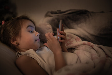 Obraz premium Cute baby girl uses phone in bed at night instead of sleeping close up.