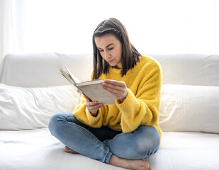 A cute girl in a yellow sweater is reading a book at home on the couch.