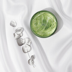 Beauty products, round jar with transparent gel and eucalyptus branch painted in silver color on white satin fabric