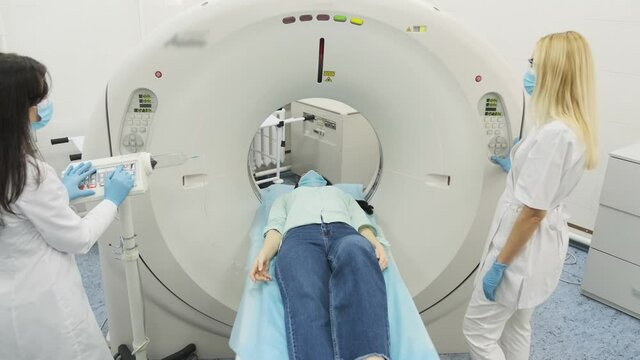 Female patient is undergoing CT or MRI scan under supervision of two qualified radiologists in mask and gloves in modern medical clinic. Patient lying on a CT or MRI scan table