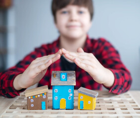 A boy in a blurr with a smile holds his hands over the wooden houses. The concept of real estate protection.