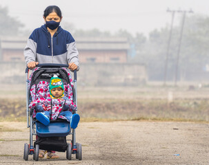 an elderly indian lady wearing black face mask pushing an indian baby in winter wear in a stroller perambulator on a road with copy space and background blur