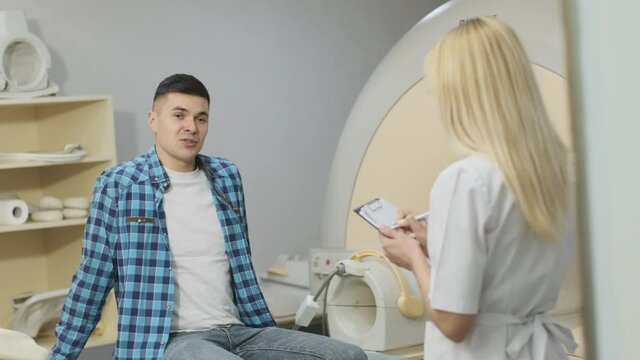 Woman doctor radiologist has a dialogue with young male patient, asks and notes the patient's medical history, complaints in modern clinic beside modern closed-type MRI