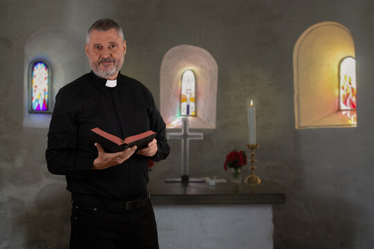 A priest in a black shirt with a white clerical collar stands in front of the altar of a small round church on the island of Bornholm. The older man has gray hair and a beard and is holding a Bible.