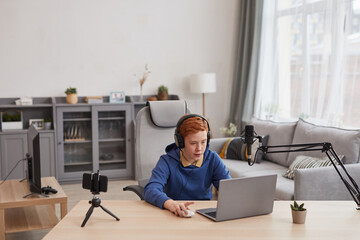 Wide angle portrait of red haired teenage boy playing video games with microphone and camera set up for online streaming, copy space