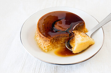 Close-up of a homemade custard pudding on a white plate and some cinnamon sticks, with a spoon ready to eat.