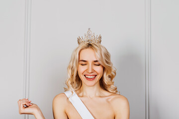 Happy oung blonde woman wearing a diamond golden crown and a white ribbon, with eyes closed, smiling. Beauty contest winner concept.