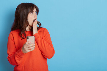 Portrait of beautiful Caucasian woman shocking with something on mobile phone, looking aside and covering opened mouth, holds mobile phone, copy space for advertisement.