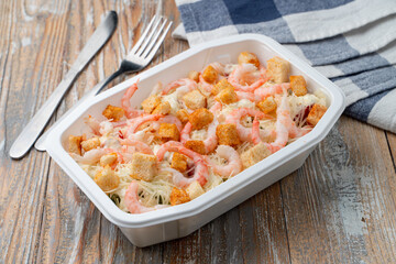 Food delivery: seafood salad with shrimps and croutons in a plastic box on light wooden table