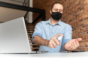 An male office employee wearing mask is spraying antibacterial sanitizer for for disinfecting hands before using laptop. Social distance and safety at work during pandemic and viral disease