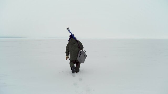 
a fisherman in a hat with earflaps walks with equipment on a frozen lake for winter fishing. 
the camera follows behind.