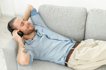 Cheerful handsome guy using wireless headphones for listening music, a man lays on the couch with eyes closed lost in dreams, enjoying favorite tracks, rest in cozy atmosphere