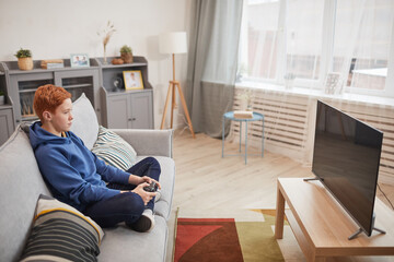 Wide angle portrait of red haired teenage boy playing video games while sitting on sofa and holding gamepad, copy space