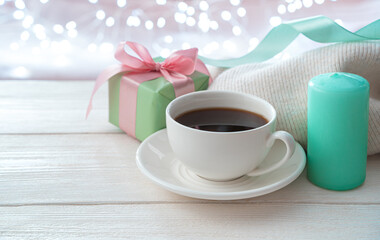 Festive background with a white ceramic coffee cup, a gift and a candle on a light background with a burning garland. Side view with copy space. The concept of holidays, March 8.