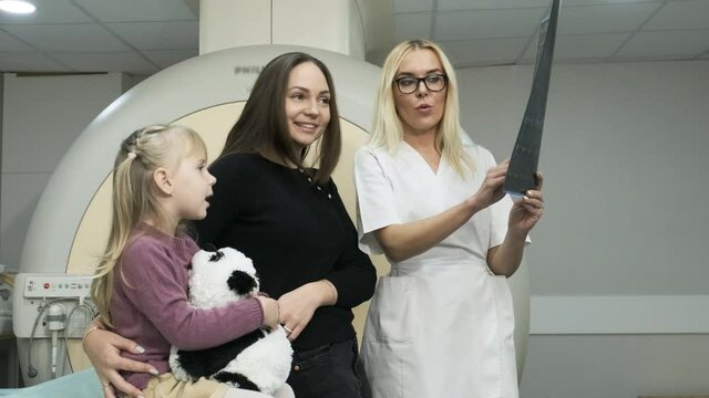 Woman doctor radiologist explains results of MRI scanning for woman with her child, showing the snapshot with images, observing and analyzing mri scan in modern clinic beside modern closed-type MRI