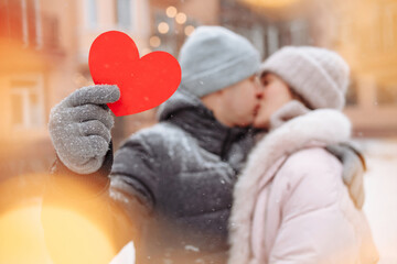 Valentine's day concept, Loving couple kiss and hug at a winter snowy park. Young man holds a red paper heart while celebrating all lover's day with his girlfriend. A couple feeling warm together.