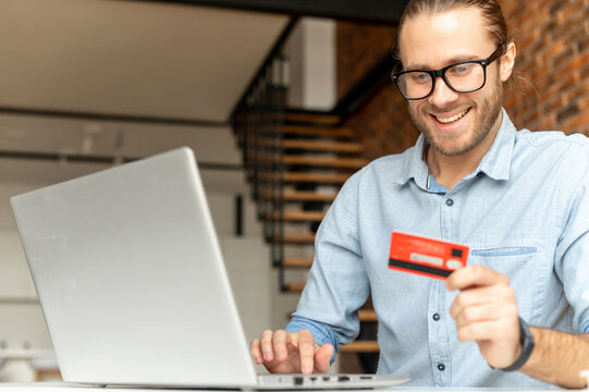A guy is makes an order online, e-shop, smiles in anticipation of the purchase, a man holding credit card, typing number data on laptop for paymant online, check balance, mobile banking, tranfer funds