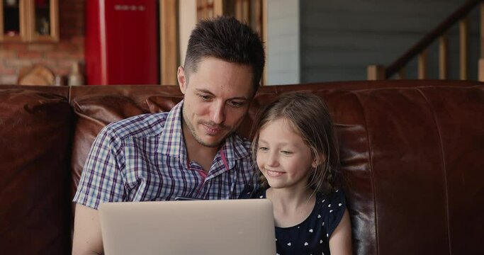 Cheerful father and little 7s daughter sit on couch spend time together at home having fun using laptop, watch funny videos on internet, cartoons on-line, modern tech usage, parental control concept