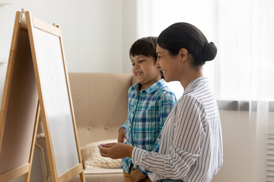 Friendly indian woman psychologist and small boy stand by easel board hold crayon planning picture talk on visual art therapy session. Happy hindu mother teach little son to draw using piece of chalk
