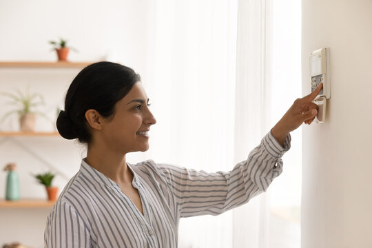 Happy Millennial Indian Female Homeowner Managing Smart House Electronic System Pressing Button At Control Panel. Young Mixed Race Woman Confident In Easy Programming Of Modern Climate Control Device