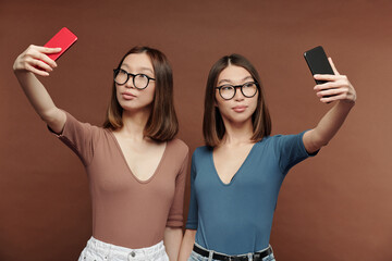 Cute twin sisters in casualwear standing close to one another and making selfie