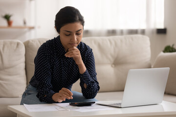 Thoughtful millennial indian lady sit on sofa engaged in paperwork at home calculate expenses planning budget. Young mixed race female homeowner count payable amount before paying utility bills online