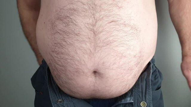 Overweight man measures his waistline. He is shirtless and his trousers are undone.