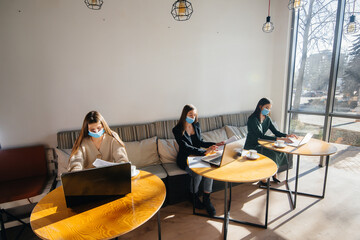 A group of masked girls keep a social distance in a cafe when working on laptops.