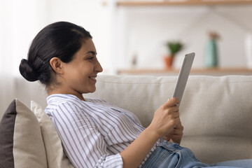 Side view of happy indian woman recline on pillows at sofa engaged in reading digital book from tablet screen. Serene mixed race young lady spend leisure time alone watch funny video on pad pc device