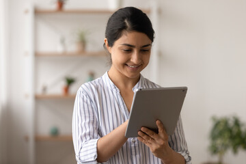 Young indian female in good mood stand at living room with tablet pc device corresponding with friend at social network. Confident smiling mixed race lady check mail client on pad using wifi internet