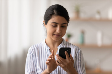 Fototapeta premium Content young indian lady look at screen of modern cellphone device check email chat at network messenger. Pleasant millennial mixed race woman use mobile phone at home browse news in convenient app