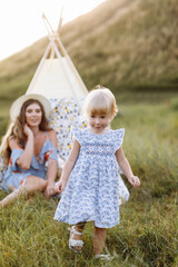 Beautiful mother with her little daughter sitting near wigwam in the field. Spending time together, outside, on vacation, outdoors. Beautiful sunset light in the garden or in the park. © Andriy Medvediuk