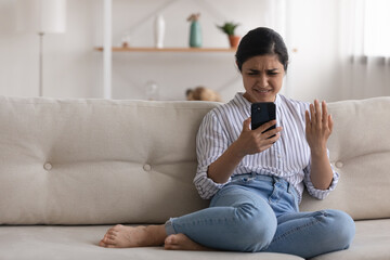 Confused angry indian woman sit on couch at home read spam message on phone screen have problem...