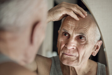 Mature hoary man looks in mirror and examined his gray hair.