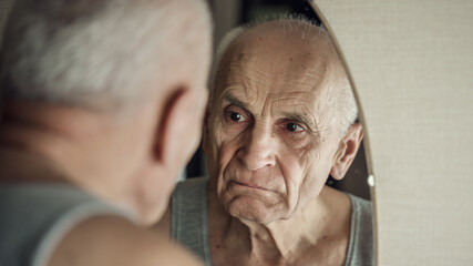 Sullen aged gray haired man looks in mirror in his face reflection