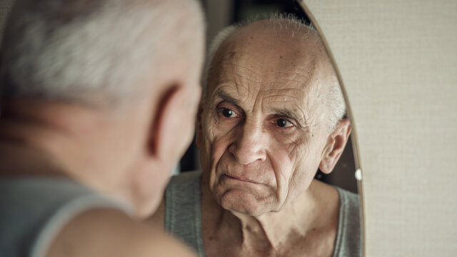 Sullen aged gray haired man looks in mirror in his face reflection