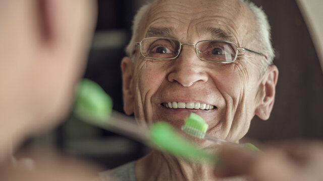 Gray haired senior man is cleaning his teeth and smiling looking in mirror