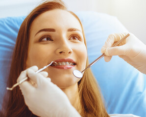 Smiling young woman with orthodontic brackets examined by dentist in sunny dental clinic. Healthy...