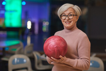 Fototapeta premium Waist up portrait of smiling senior woman holding bowling ball and looking at camera while enjoying active entertainment at bowling alley, copy space