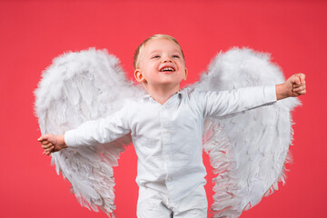 Obraz premium Excited angel little boy with white wings. Happy angelic children boy laughing. Cute kid with white wings. Adorable child cupid isolated on red. Valentines day card, baner copy spase. Funny face.
