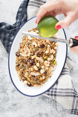 Woman hands grating a lime over a metallic white tray full of delicious granola over a rustic napkin on a white and grey background.